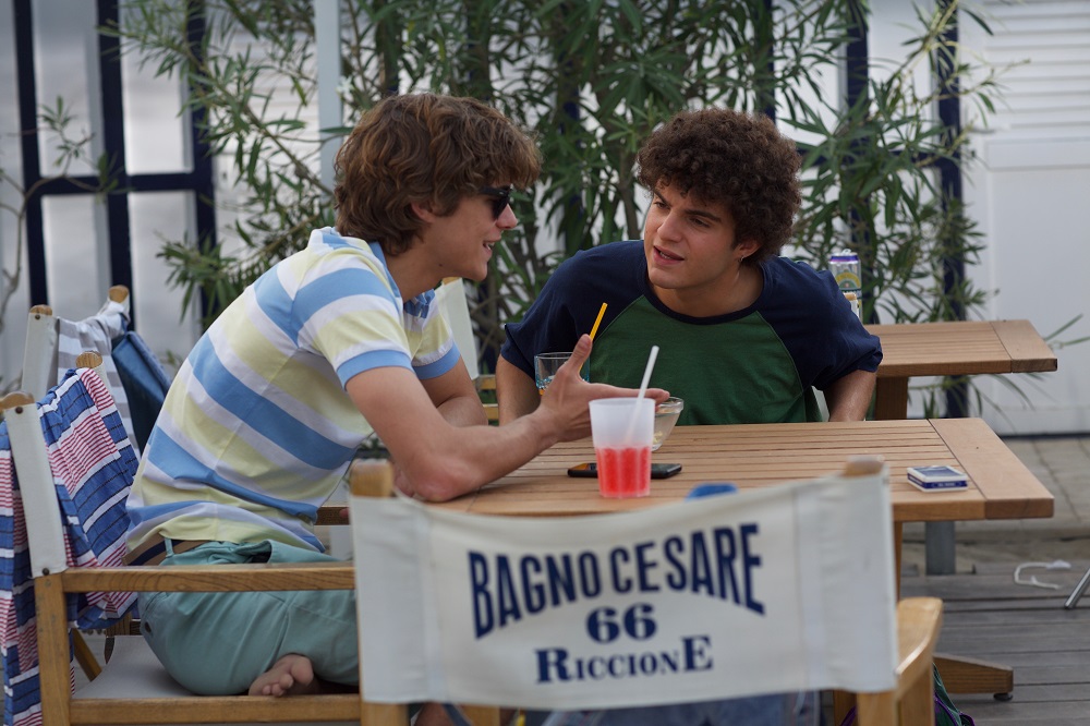 Lorenzo Zurzolo (Vincenzo) e Davide Calgaro (Furio) in una scena di Sotto il sole di Riccione. Sono entrambi seduti ad un tavolino del lido e stanno parlando.  